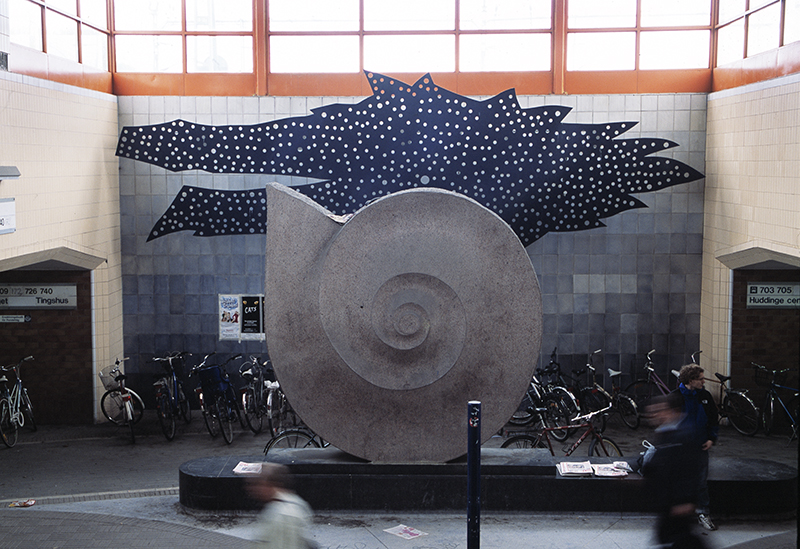 En stor snäcka av sten framför cyklar och en blå vägg av kakel. Bakom snäckan en stor blå abstrakt perforerad form.