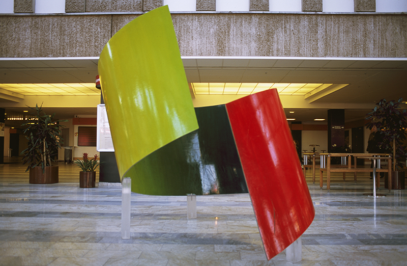 Böjd figur målad i rött och grönt som står på ett ljust golv inomhus.