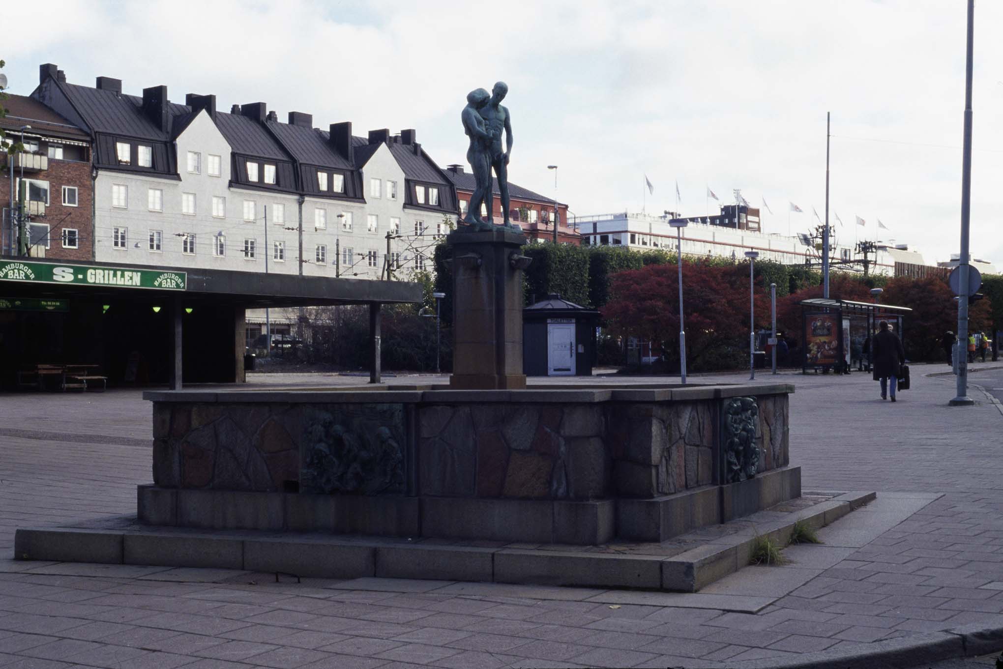 En skulptur av en man och kvinna som står på en stenpelare. Pelaren är i mitten av fyrkantig form av sten i torgmiljö.