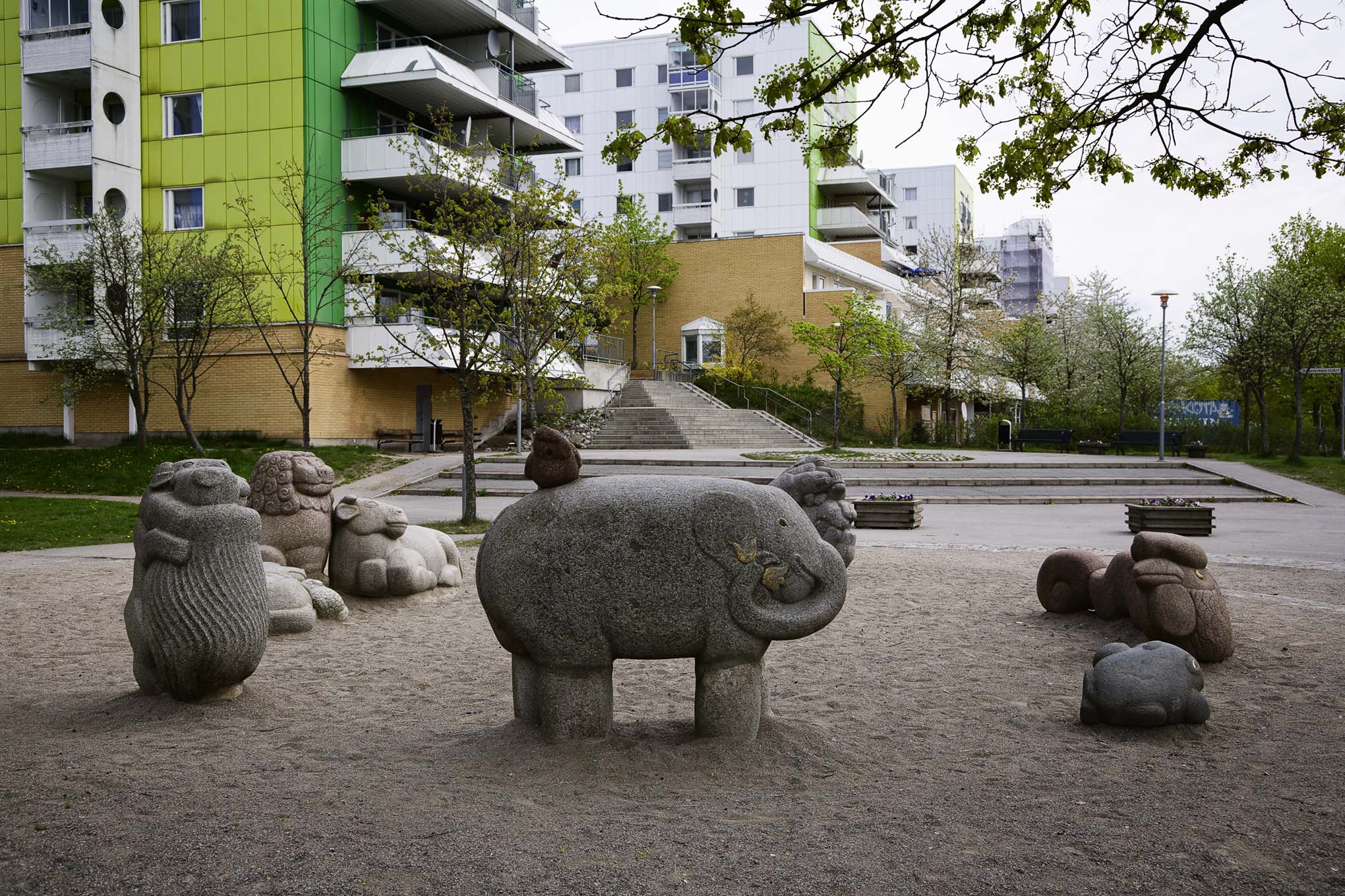 Djur gjorda av sten som står i sanden. Gröna och vita flervåningshus bakom. Grönskande träd.