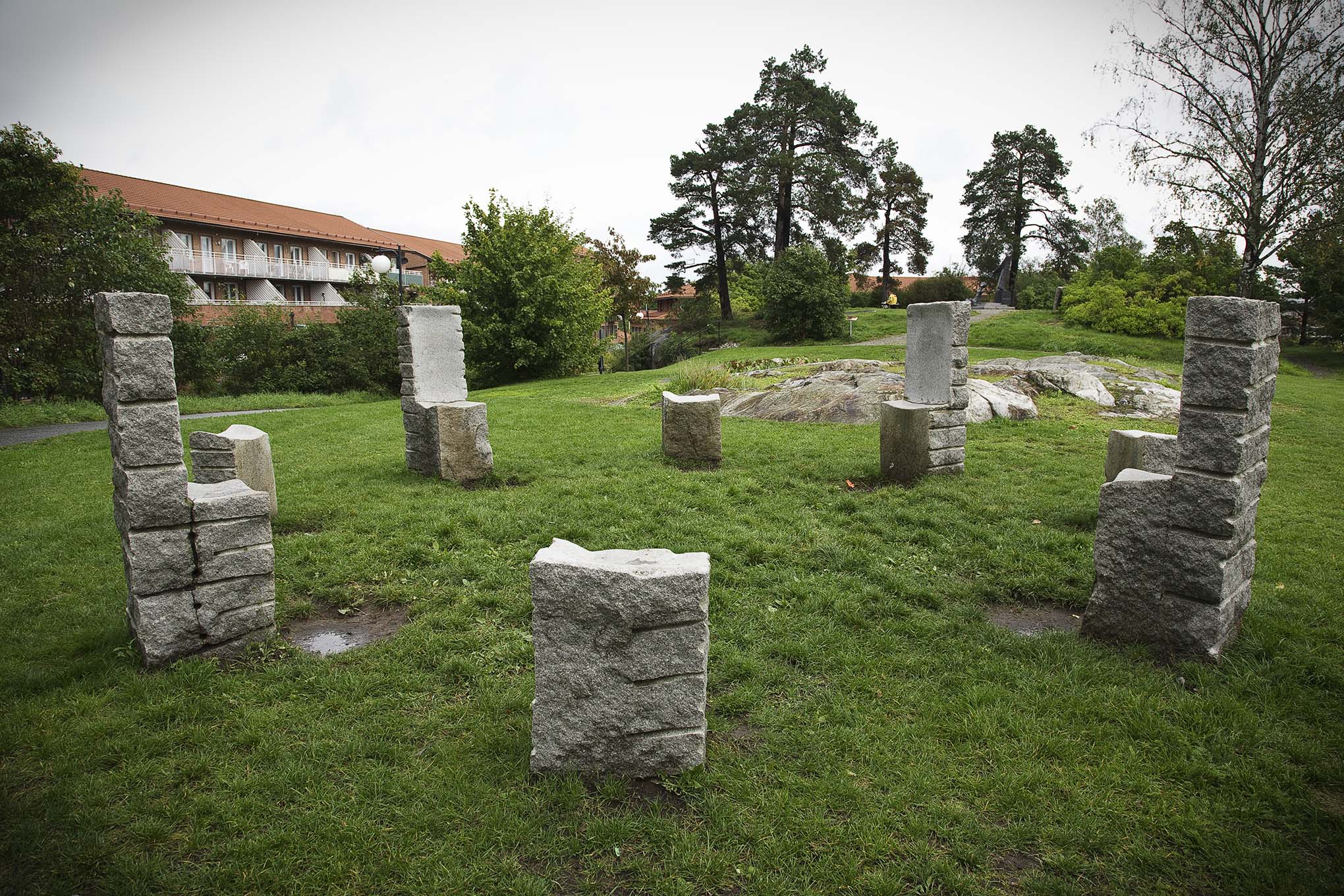 Åtta stolar av sten som står i en ring på en gräskulle. Träd och hus bakom.