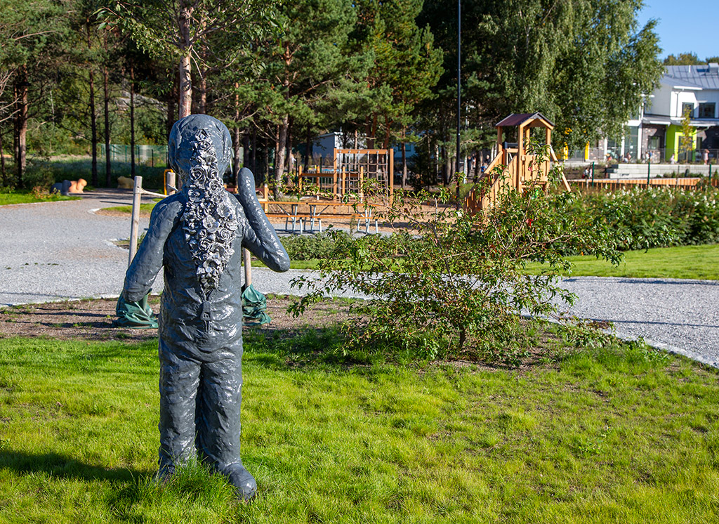 En grå skulptur som ser ut som en människa men utan ansikte. Figuren har en overall som är uppknäppt och där finns en massa blommor.