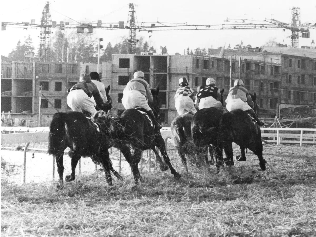 Svartvit bild av en klunga med hästar och ryttare. I bakgrunden byggs ett bostadsområde.