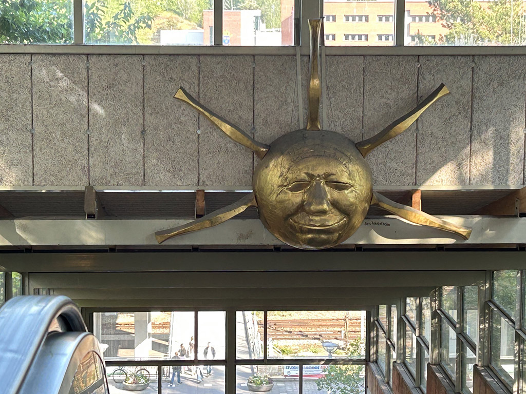 En guldfärgad sol med fem strålar som blundar och ler på väggen ovanför en rulltrappa med ett fönster ovanför. Hus och grönska utanför.